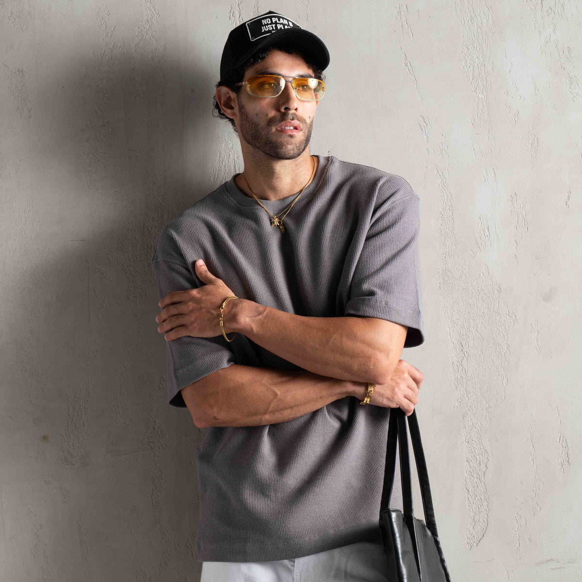A man wearing a dark gray t-shirt, black baseball cap, and yellow-tinted sunglasses leans against a textured gray wall, holding a black tote bag.