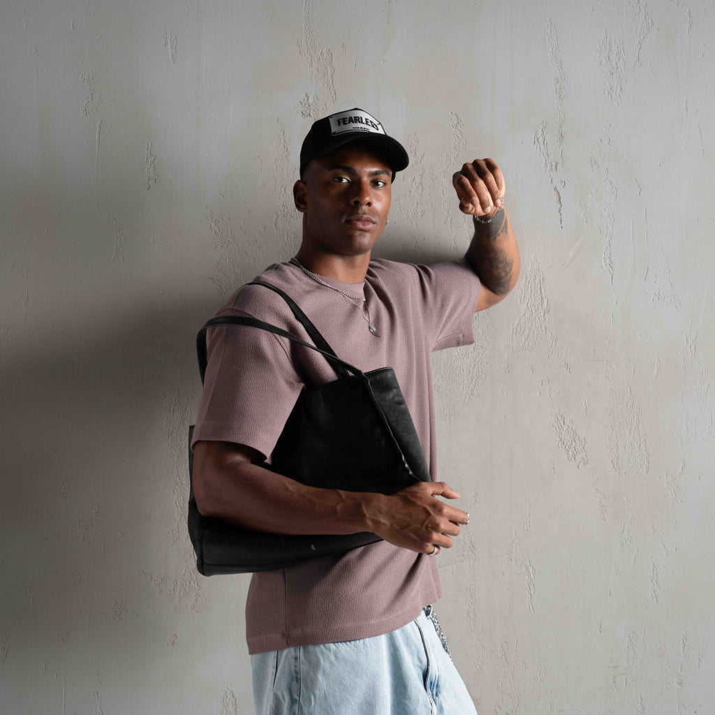 A man wearing a dusty mauve t-shirt, light blue jeans, and a black baseball cap with a white logo, carrying a black leather shoulder bag against a textured gray wall.